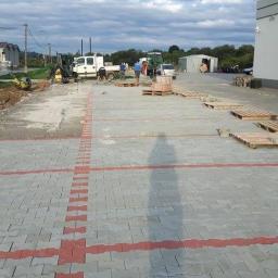 Damian Mazur - Fragment powierzchni z ułożoną szarą kostką brukową z czerwonymi akcentami, palety z kostką i ekipa robotników w tle, widok z góry z cieniem osoby.