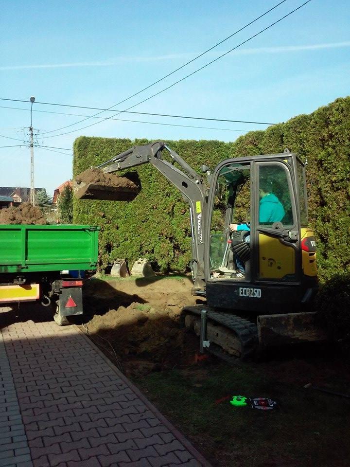 Mini koparka Volvo ECR25D załadowuje ziemię do zielonej przyczepy na tle żywopłotu i kostki brukowej.