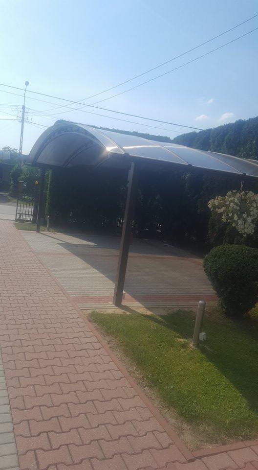 Zadaszenie nad miejscem parkingowym, wykonane z przezroczystego tworzywa sztucznego, wsparte na metalowych słupach, przylegające do chodnika z czerwonej kostki brukowej.