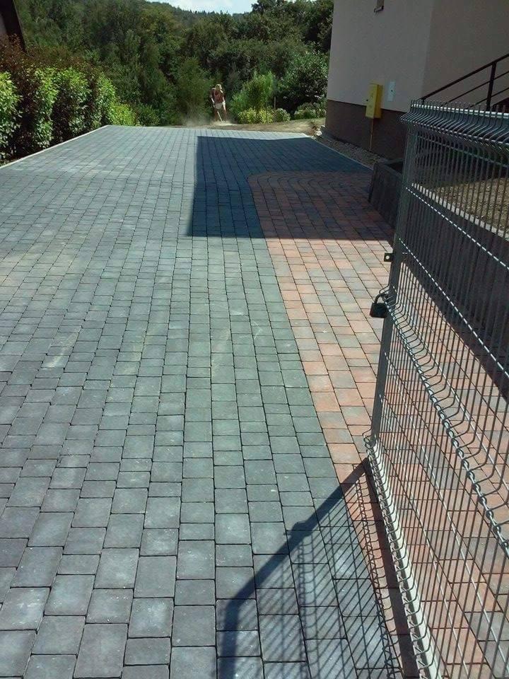 Ułożona szara kostka brukowa na podjeździe, z elementami z czerwonej kostki przy budynku. Widoczna brama z metalowej siatki i osoba w oddali, prawdopodobnie sprzątająca.