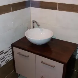 Umywalka nablatowa w kształcie misy, chromowana bateria, szafka łazienkowa z szufladami, płytki ceramiczne w odcieniach bieli i brązu z dekoracyjnym pasem, fragment brązowych drzwi z klamką.