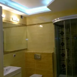 Wykończona łazienka z beżowymi płytkami, podświetlanym sufitem i kabiną prysznicową z geometrycznym wzorem na szkle.