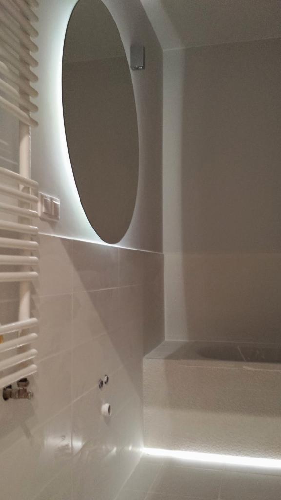 Nowoczesna łazienka z podświetlanym owalnym lustrem, grzejnikiem drabinkowym i subtelnym oświetleniem LED pod wanną.