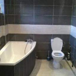 Wykończona łazienka z wanną, toaletą i grzejnikiem drabinkowym. Ściany wyłożone ciemnymi płytkami z poziomym pasem jasnych płytek. Widoczny uchwyt na papier toaletowy i ręczniki na grzejniku.