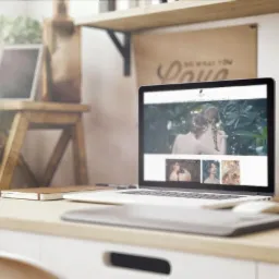 Laptop wyświetlający stronę internetową z artystycznymi zdjęciami, umieszczony na biurku z roślinami i dekoracjami w tle.