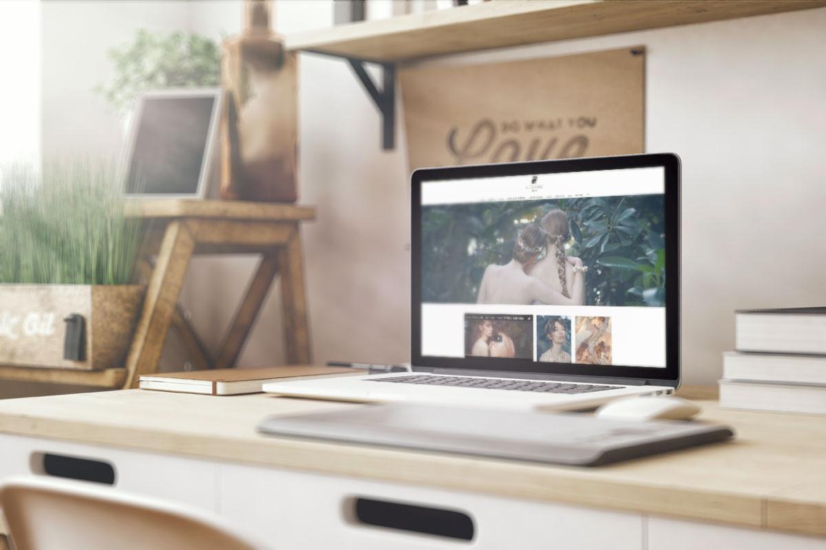 Laptop wyświetlający stronę internetową z artystycznymi zdjęciami, umieszczony na biurku z roślinami i dekoracjami w tle.