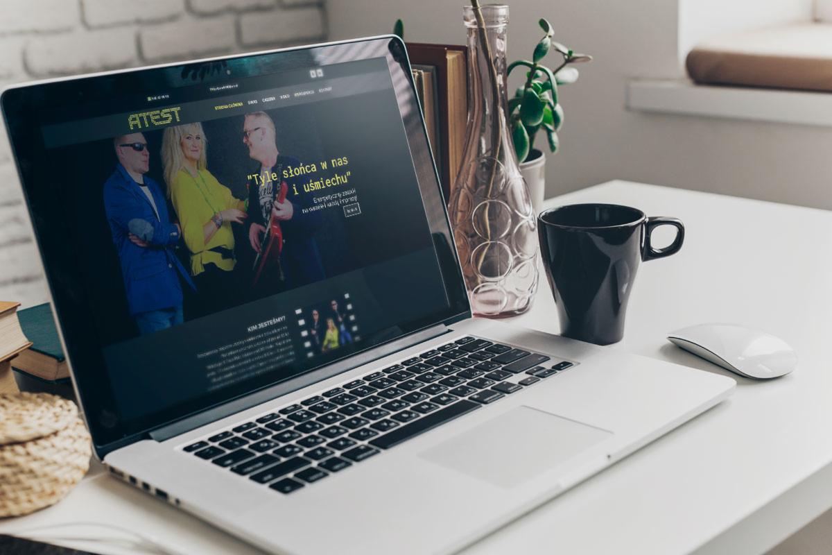 Laptop wyświetlający stronę zespołu muzycznego 'ATEST' na biurku z czarnym kubkiem, myszką i wazonem, obok książki i pleciony koszyk.