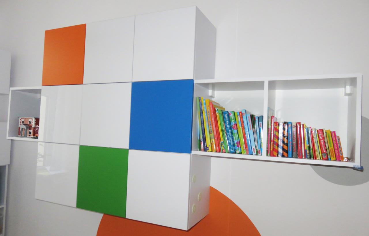 Białe, modułowe szafki wiszące z kolorowymi frontami (pomarańczowy, niebieski, zielony) i otwartą półką z książkami dla dzieci oraz dekoracyjnym modelem domu w środku.
