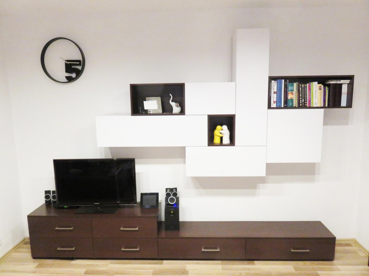 Nowoczesna aranżacja ściany w salonie z brązową szafką RTV, telewizorem, głośnikami i białymi, modułowymi półkami z dekoracjami i książkami, uzupełniona o minimalistyczny zegar.
