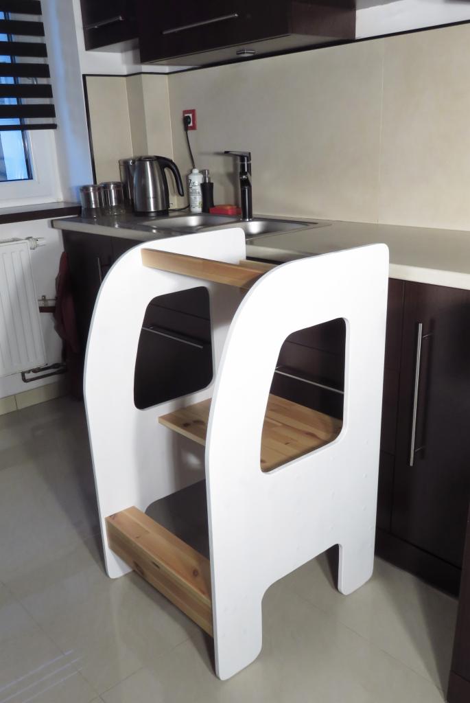 Biały, drewniany podest dla dziecka w kuchni, umożliwiający dostęp do blatu, z widocznymi półkami i zaokrąglonymi bokami, na tle zlewu i szafek kuchennych.