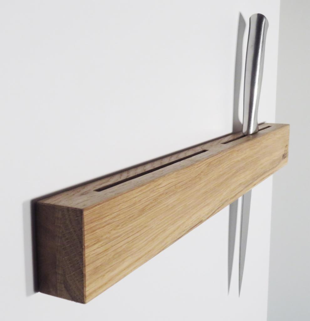Minimalistyczny uchwyt na noże wykonany z jasnego drewna dębowego, zamontowany na białej ścianie, z włożonymi dwoma nożami o srebrnych ostrzach i rękojeściach.