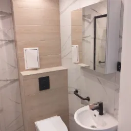 Nowoczesna łazienka z wiszącą toaletą, umywalką nablatową z czarną armaturą i lustrem z szafką, wykończona płytkami imitującymi drewno i marmur.