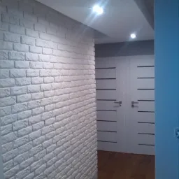 Korytarz z dekoracyjną ścianą z białej cegły, drewnianą podłogą i białymi drzwiami z poziomymi, czarnymi paskami.