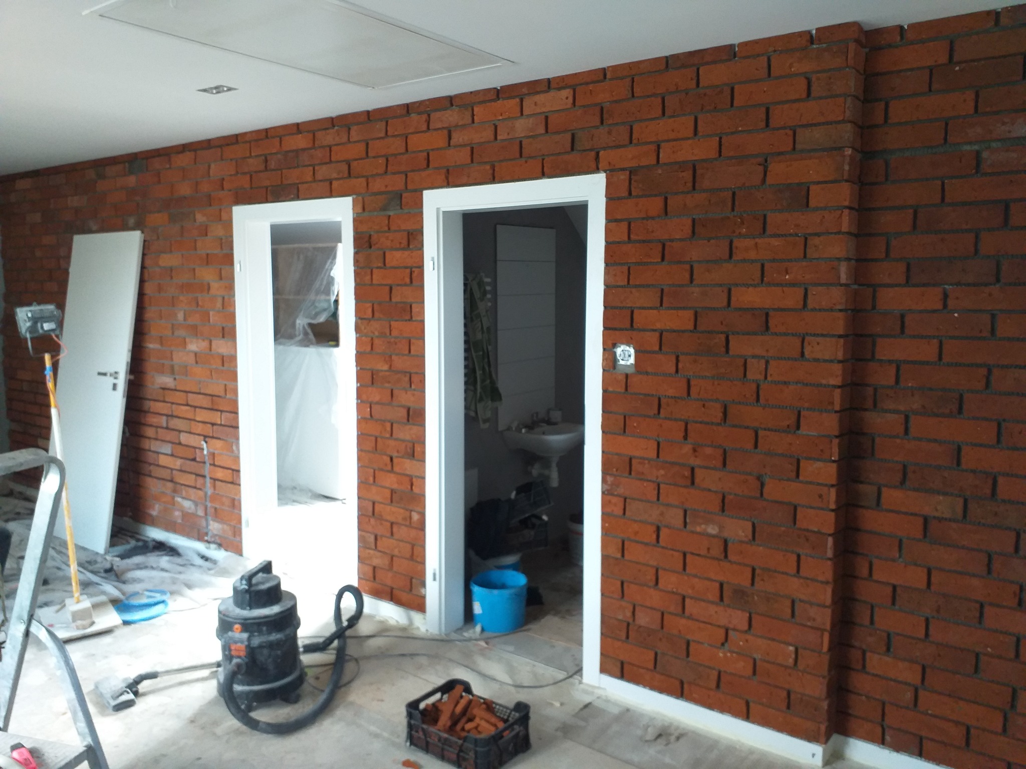 Ściana z cegły klinkierowej w trakcie prac wykończeniowych, widoczne dwie ościeżnice drzwiowe, narzędzia i akcesoria budowlane na podłodze.