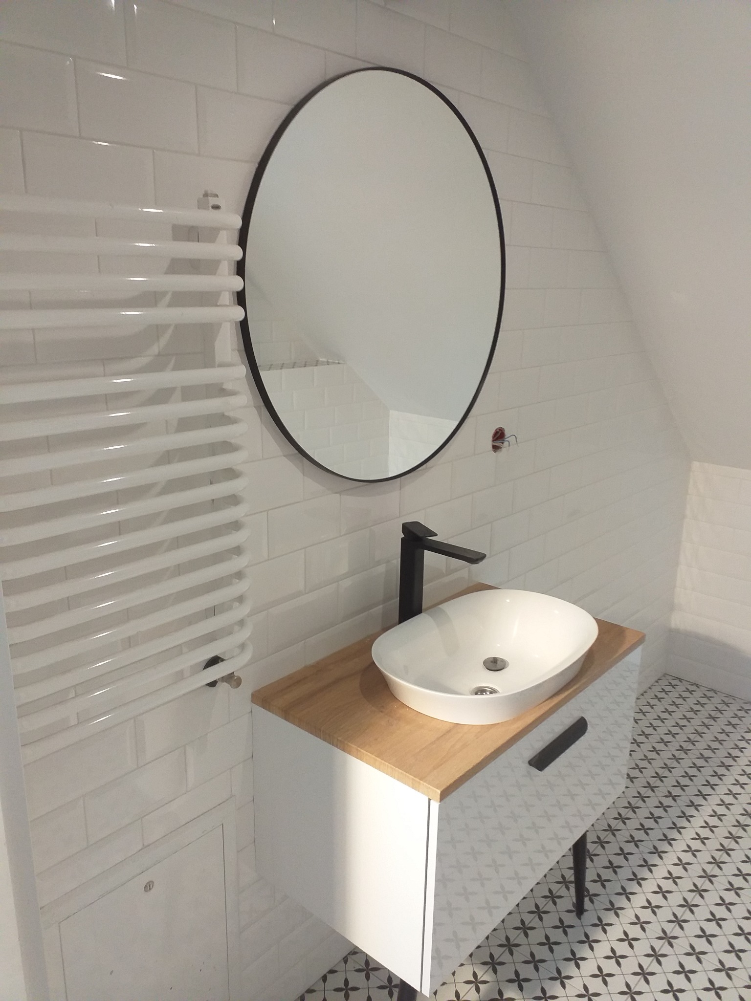 Nowoczesna łazienka na poddaszu: białe płytki, okrągłe lustro w czarnej ramie, biały grzejnik drabinkowy, umywalka nablatowa na szafce z drewnianym blatem i wzorzysta podłoga.