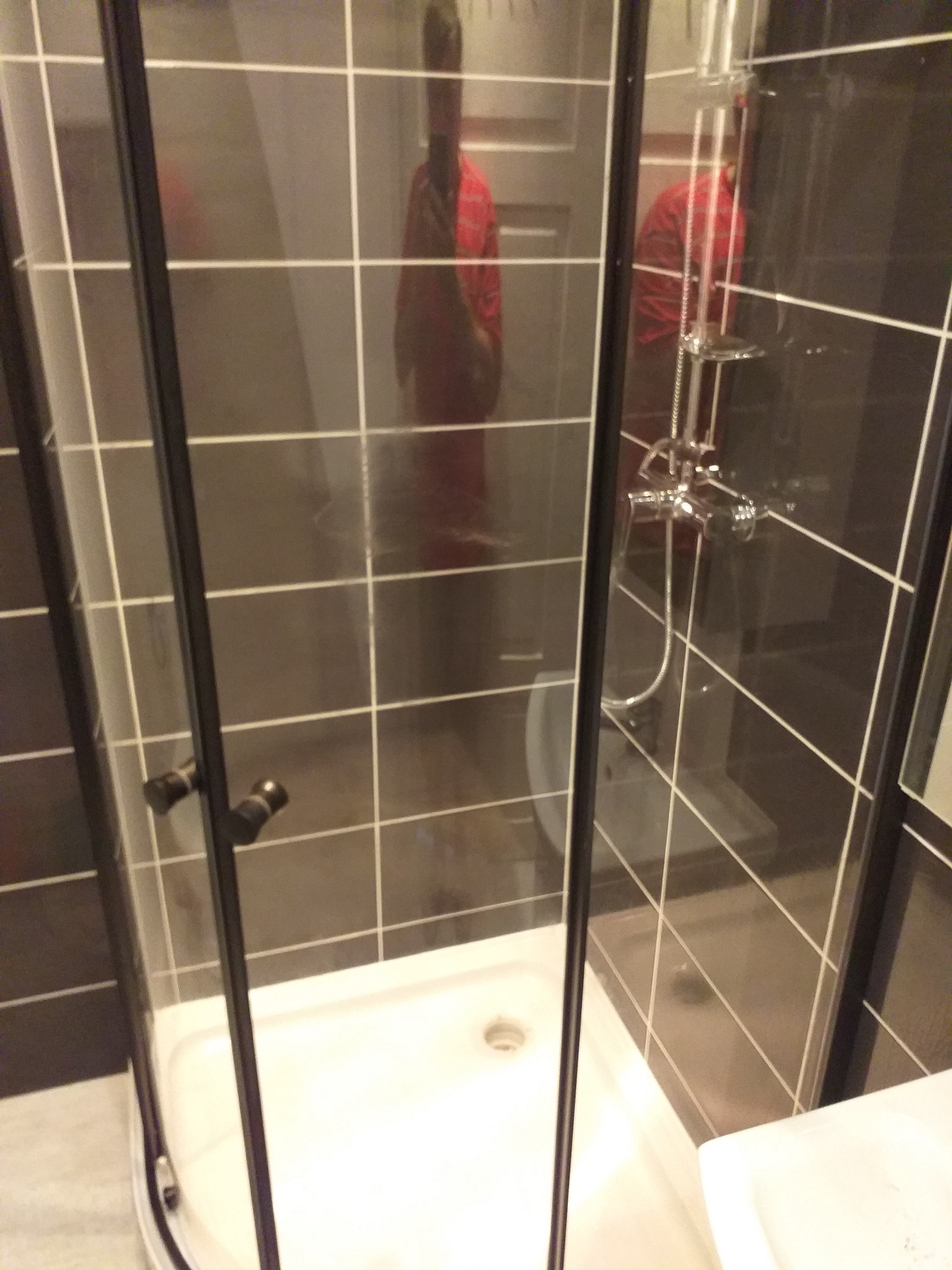 Kabina prysznicowa z czarnymi profilami i szarymi płytkami ułożonymi w kratkę, widoczna armatura i czerwona koszula wisząca na wieszaku w odbiciu.