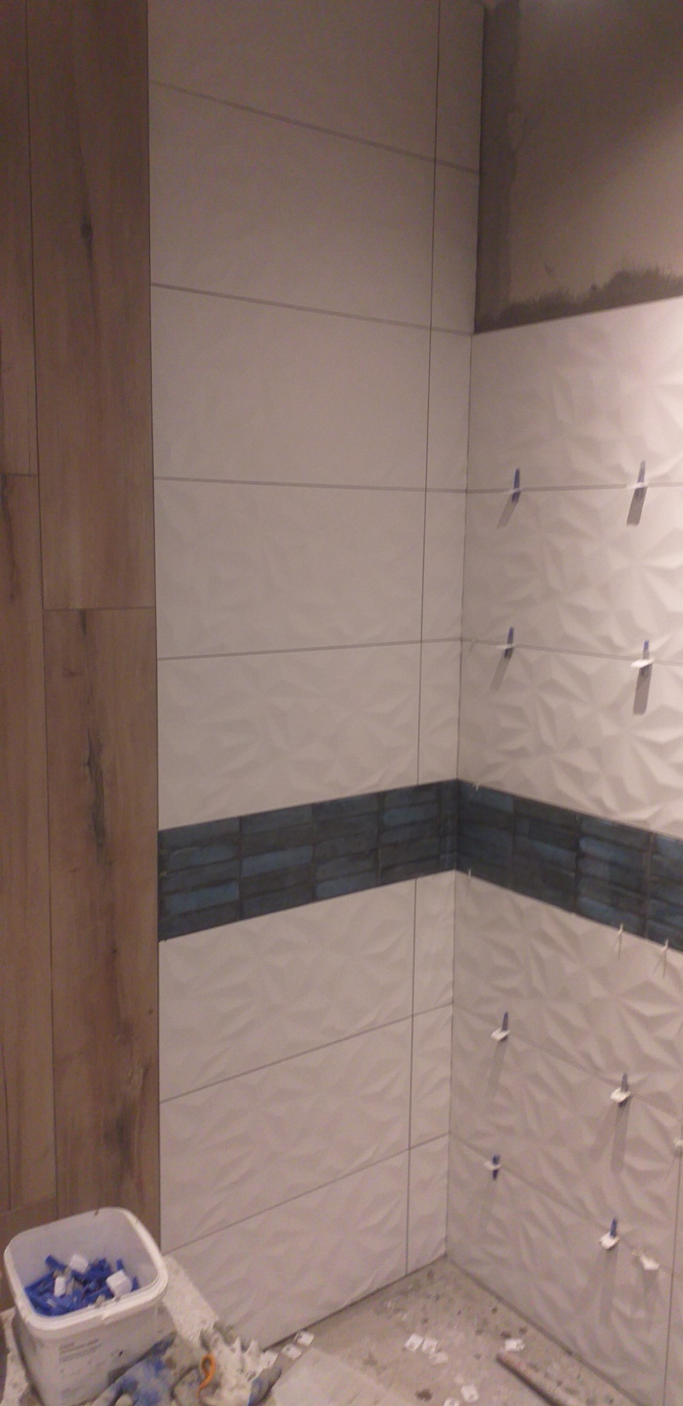 Ściana w trakcie układania płytek z dekoracyjnym pasem niebieskich płytek, widoczne krzyżyki dystansowe i wiadro z zapasem krzyżyków.