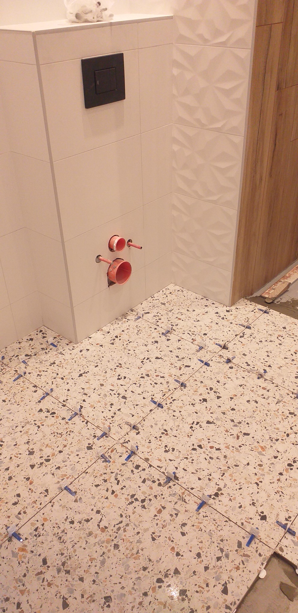 Łazienka w trakcie wykańczania: ułożone płytki podłogowe typu lastryko z niebieskimi krzyżykami dystansowymi, na ścianie białe płytki z fakturą 3D, widoczne rury kanalizacyjne i czarny przycisk...