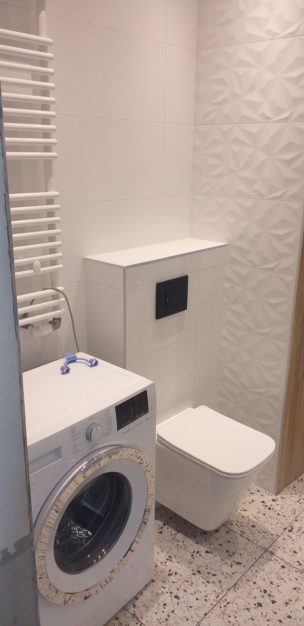 Nowoczesna łazienka z białą pralką, podwieszanym WC z czarnym przyciskiem, grzejnikiem drabinkowym i trójwymiarowymi białymi płytkami na ścianie, uzupełniona podłogą z terazzo.