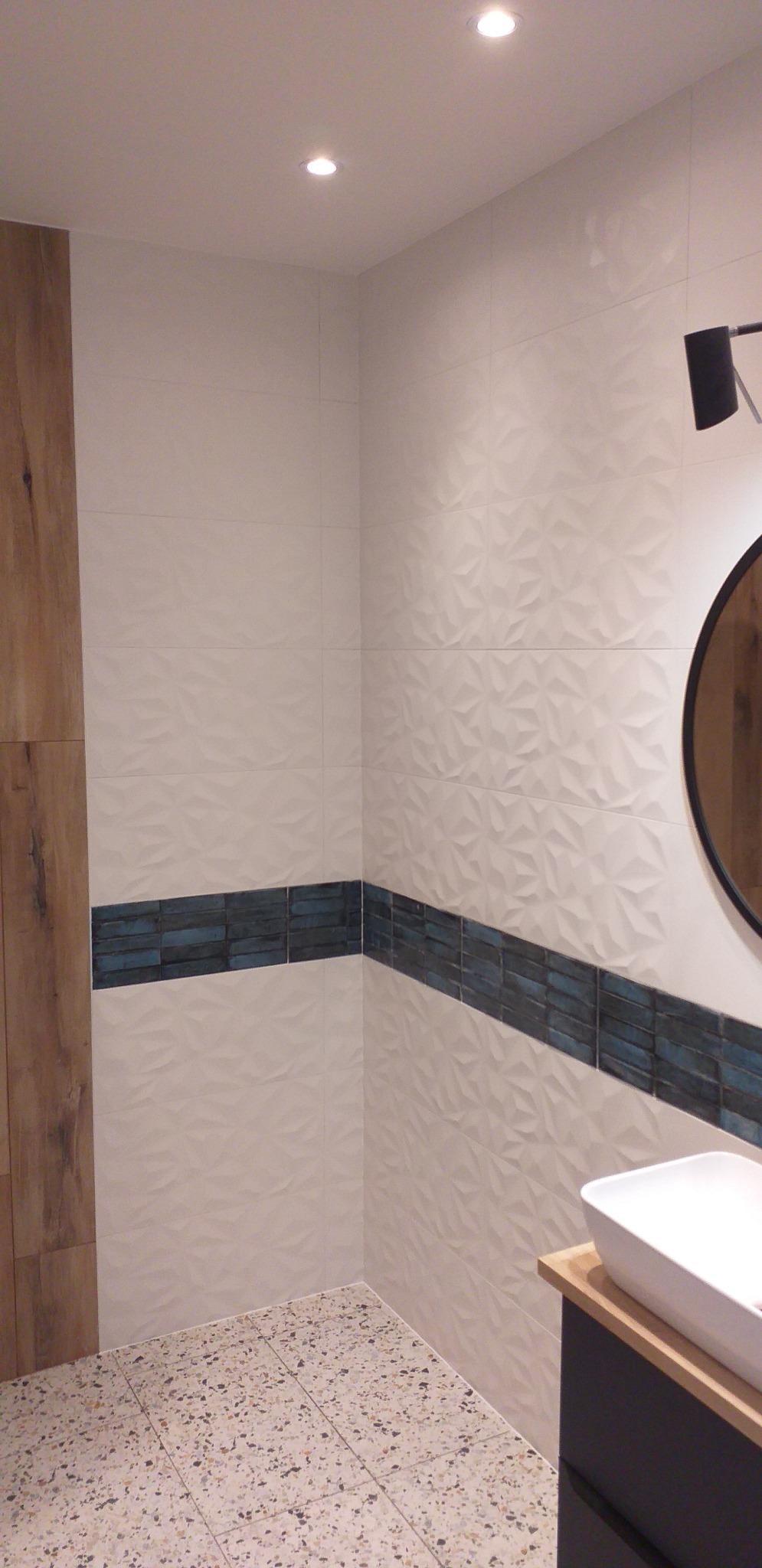 Nowoczesna łazienka z białymi trójwymiarowymi płytkami, poziomym pasem dekoracyjnym z niebieskich płytek, podłogą z terazzo i okrągłym lustrem z czarną ramą.