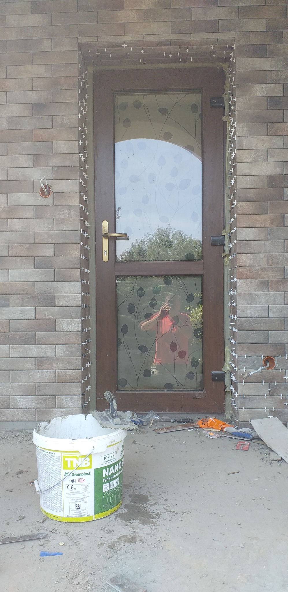 Montaż cegiełek klinkierowych wokół brązowych drzwi wejściowych z szybą z wzorem roślinnym, widoczne krzyżyki dystansowe, wiadro z zaprawą klejową i odbicie fotografa w szybie.