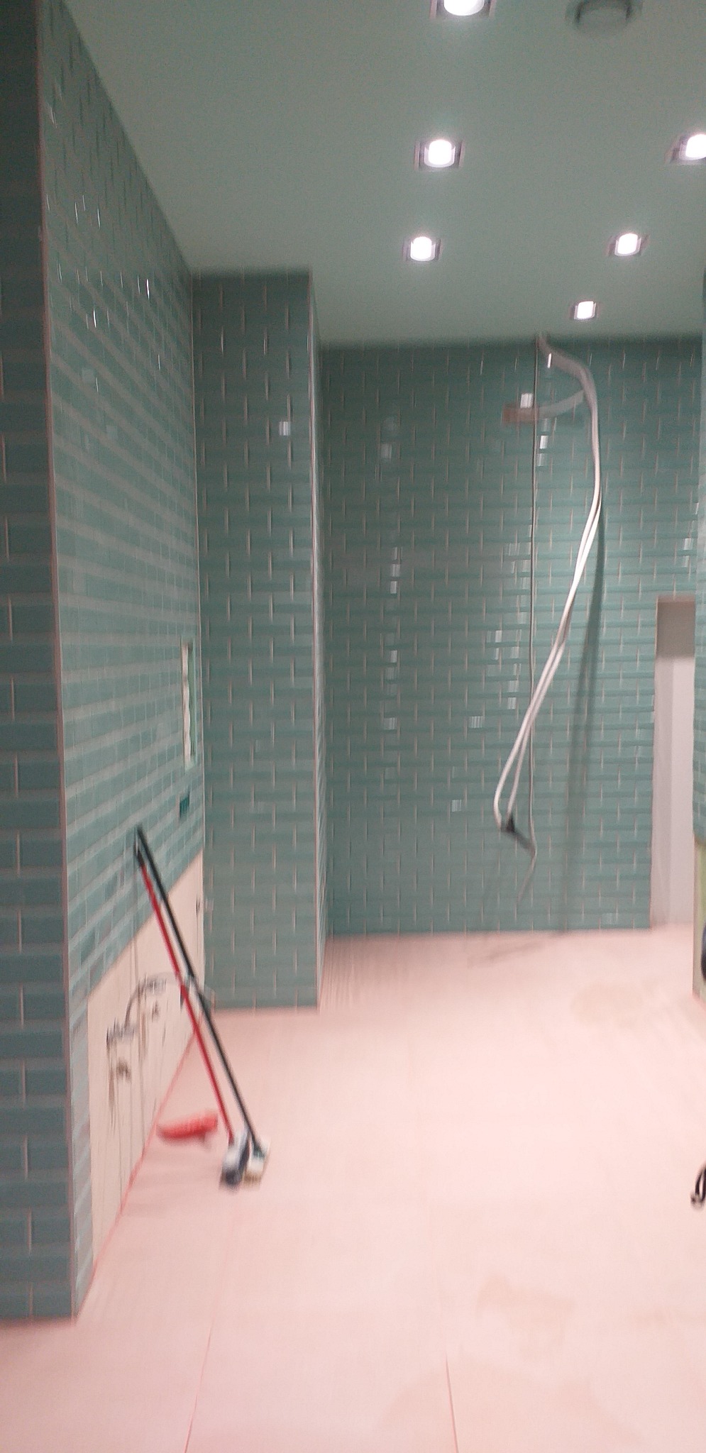 Łazienka w trakcie remontu z turkusową cegiełkową glazurą na ścianach, widoczne narzędzia glazurnicze oparte o ścianę i wystające kable od prysznica.