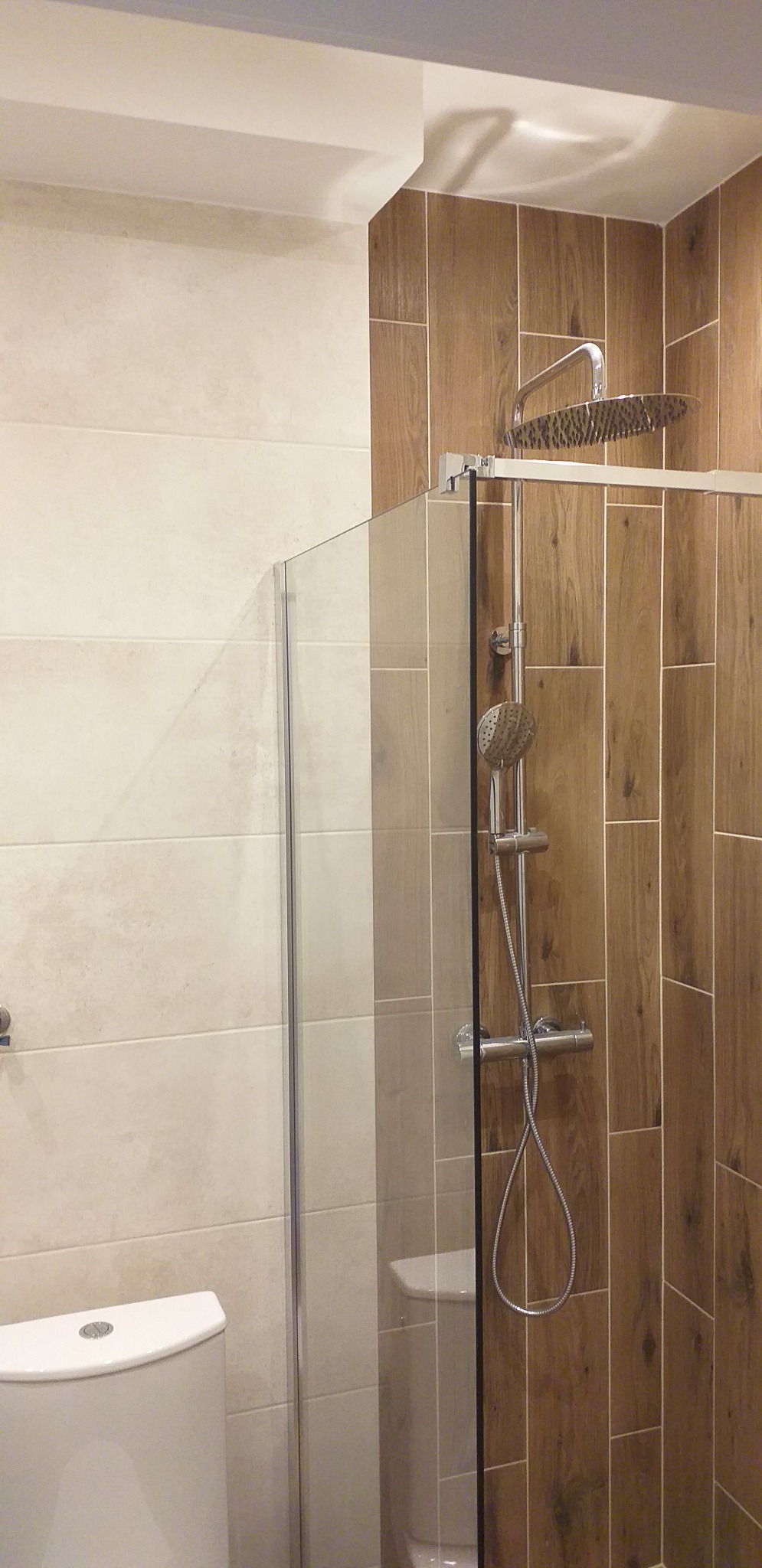 Kabina prysznicowa typu walk-in z deszczownicą i słuchawką, wykończona płytkami imitującymi drewno na jednej ścianie i jasnymi płytkami na drugiej, widoczny fragment toalety.