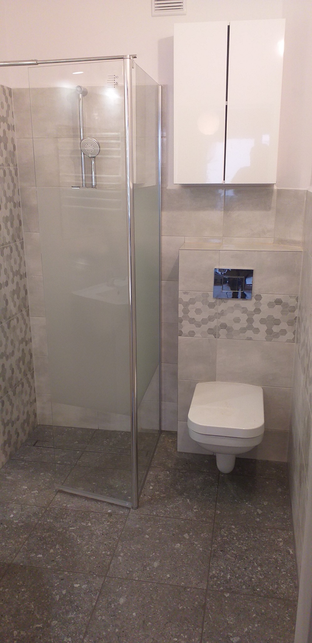 Nowoczesna łazienka z kabiną prysznicową ze szkła hartowanego, podwieszaną toaletą i białymi szafkami, wykończona szarą płytką z geometrycznym wzorem w strefie WC.