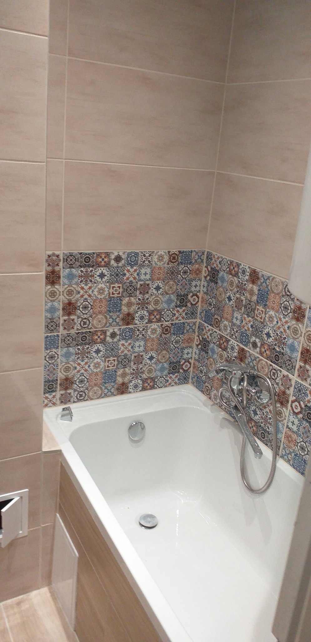 Wykończona łazienka z wanną, beżowymi płytkami i dekoracyjnym pasem mozaiki nad wanną, widoczna bateria wannowa z wężem.