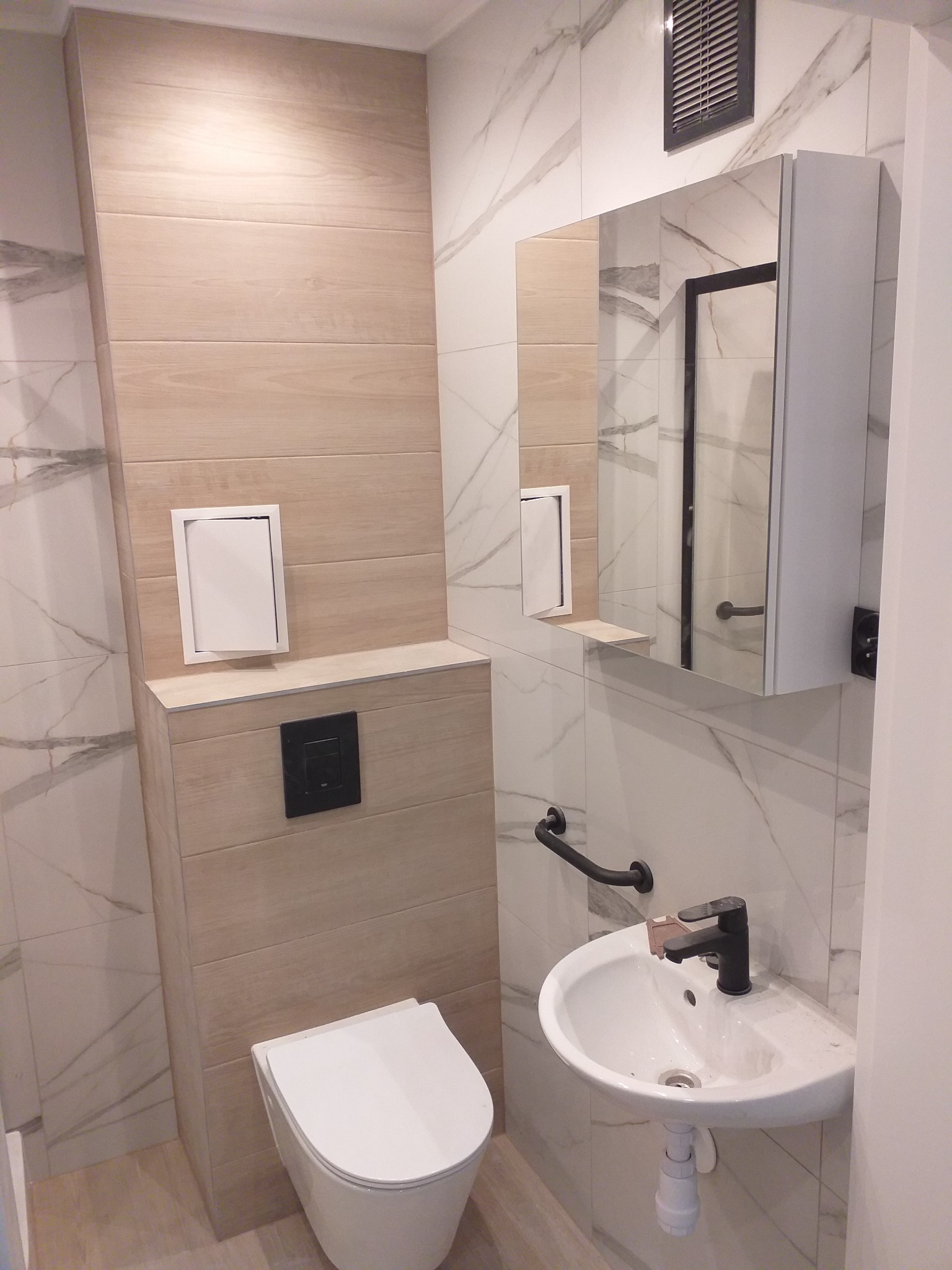 Nowoczesna łazienka z wiszącą toaletą, umywalką nablatową z czarną armaturą i lustrem z szafką, wykończona płytkami imitującymi drewno i marmur.