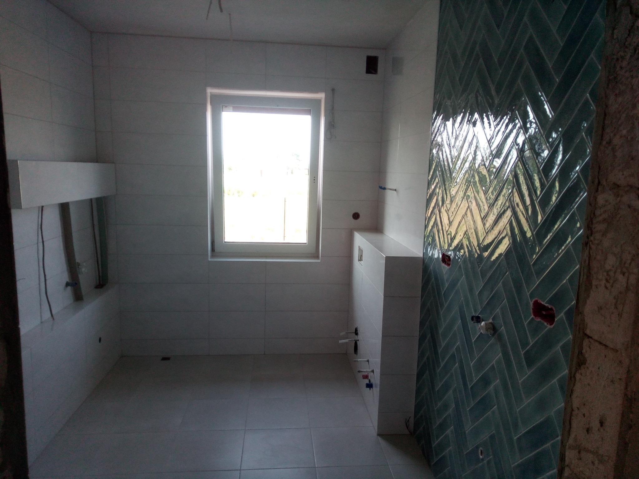 Wnętrze łazienki w trakcie prac wykończeniowych: ściany i podłoga wyłożone jasnymi płytkami, fragment ściany ozdobiony ciemnymi, błyszczącymi płytkami ułożonymi w jodełkę, widoczne instalacje...