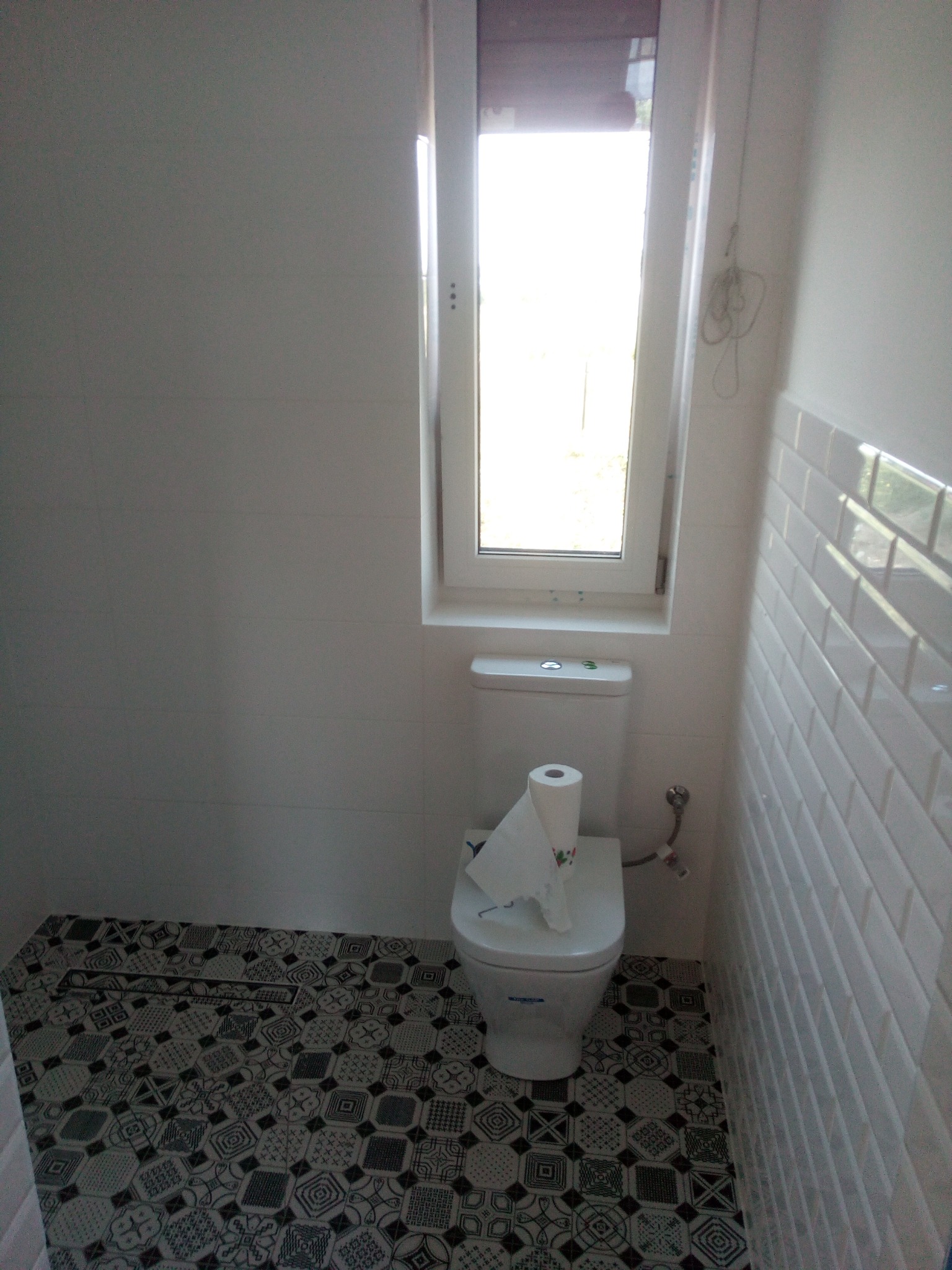 Wykończona łazienka z białą armaturą, wzorzystą podłogą i białymi, prostokątnymi płytkami na ścianie, oświetlona przez okno.