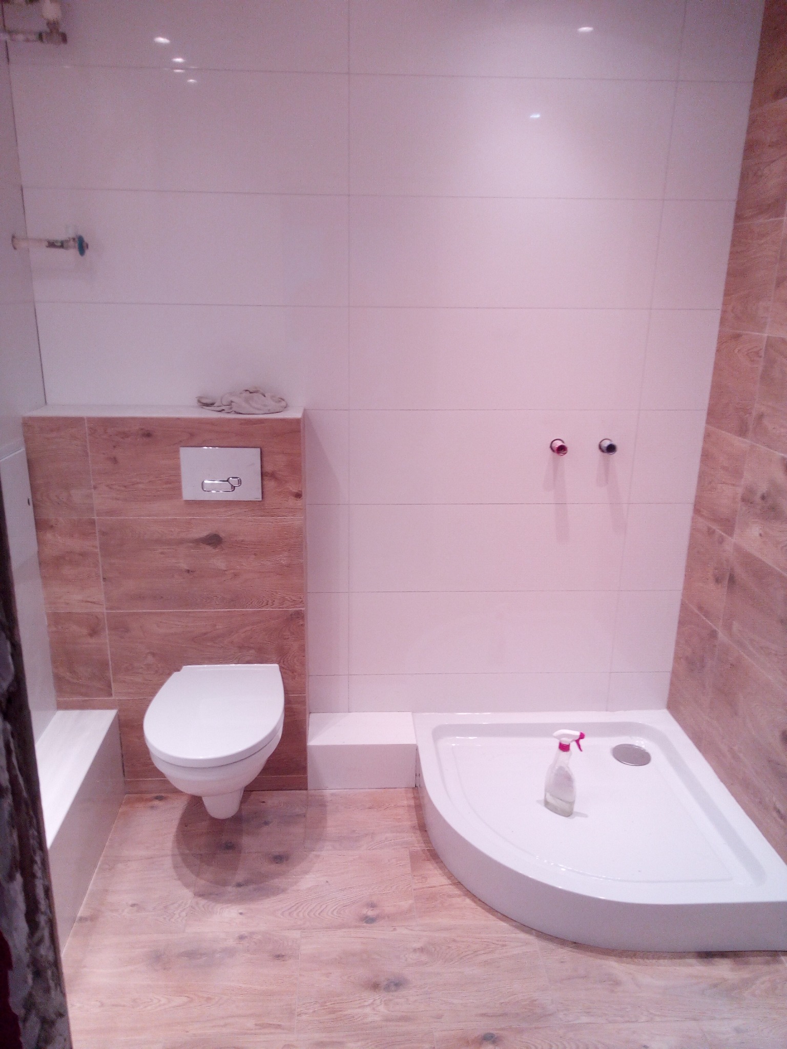 Nowoczesna łazienka z płytkami imitującymi drewno na podłodze i obudowie toalety, białe płytki na ścianach, kabina prysznicowa z półokrągłym brodzikiem, wisząca toaleta i butelka ze spryskiwaczem...