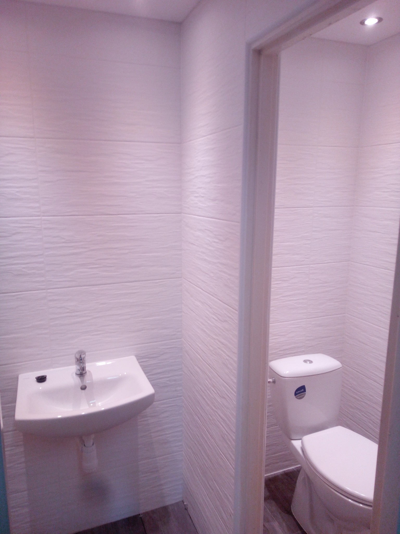 Biała umywalka i toaleta w małej łazience wykończonej białymi płytkami o falistej strukturze, widok zza futryny drzwi.