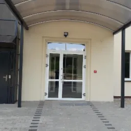 Wejście do budynku z przeszklonymi drzwiami w białej ramie, zadaszone szarą konstrukcją, z kostką brukową na podejściu i czarnymi słupami podtrzymującymi zadaszenie.