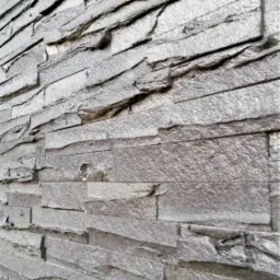 Szara ściana wykończona dekoracyjnymi płytkami przypominającymi łupki, ułożonymi warstwowo z widoczną fakturą kamienia.