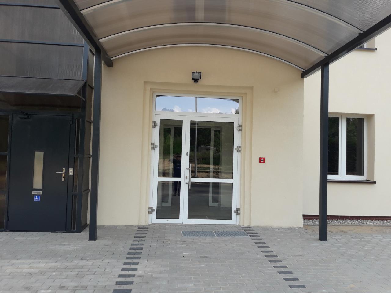 Wejście do budynku z beżową elewacją, przeszklonymi drzwiami i zadaszeniem z poliwęglanu, widoczne kostki brukowe na chodniku i okno z prawej strony.