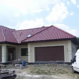 Nowo wybudowany dom jednorodzinny z czerwoną dachówką, widoczne palety budowlane i samochód dostawczy przed budynkiem.