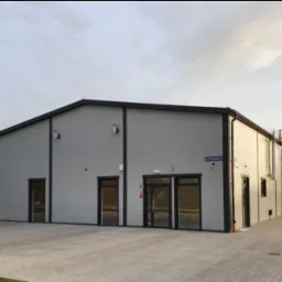 Zewnętrzny widok nowoczesnego, szarego magazynu z trzema wejściami i r&oacute;wną kostką brukową przed budynkiem. Widoczny znak z nazwą ulicy.