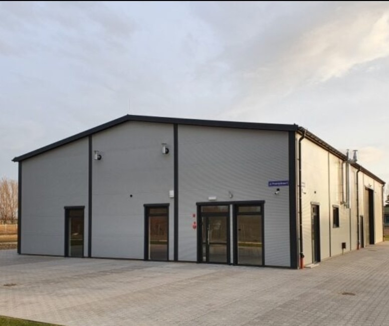 Zewnętrzny widok nowoczesnego, szarego magazynu z trzema wejściami i równą kostką brukową przed budynkiem. Widoczny znak z nazwą ulicy.