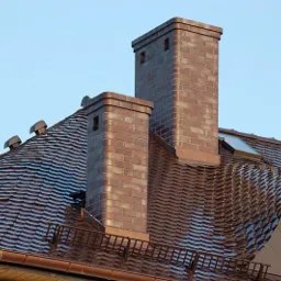 Dach pokryty dachówką karpiówką w odcieniach brązu z dwoma kominami wykończonymi cegłą klinkierową, widoczne okno dachowe i orynnowanie w kolorze miedzianym.