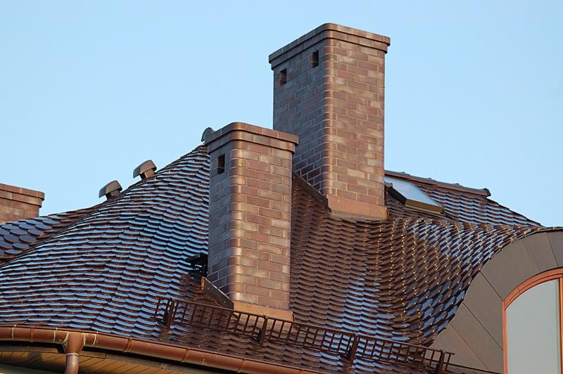 Dach pokryty dachówką karpiówką w odcieniach brązu z dwoma kominami wykończonymi cegłą klinkierową, widoczne okno dachowe i orynnowanie w kolorze miedzianym.