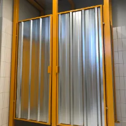 Kabina sanitarna z blachy falistej w żółtej ramie, widok z przodu, białe płytki na podłodze i ścianach.