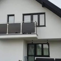 Nowoczesny dom z balkonem zabezpieczonym szklaną balustradą z metalowymi słupkami, widok z zewnątrz, jasna elewacja, czarna stolarka okienna i dach.