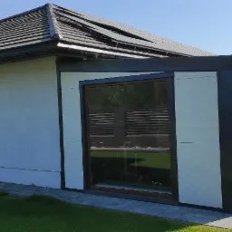 Nowoczesna, biała hala z płyt warstwowych z czarnymi obramowaniami okien i dachu, widoczna z zewnątrz w słoneczny dzień, z ułożoną kostką brukową i przygotowanymi płytami warstwowymi na gruncie.