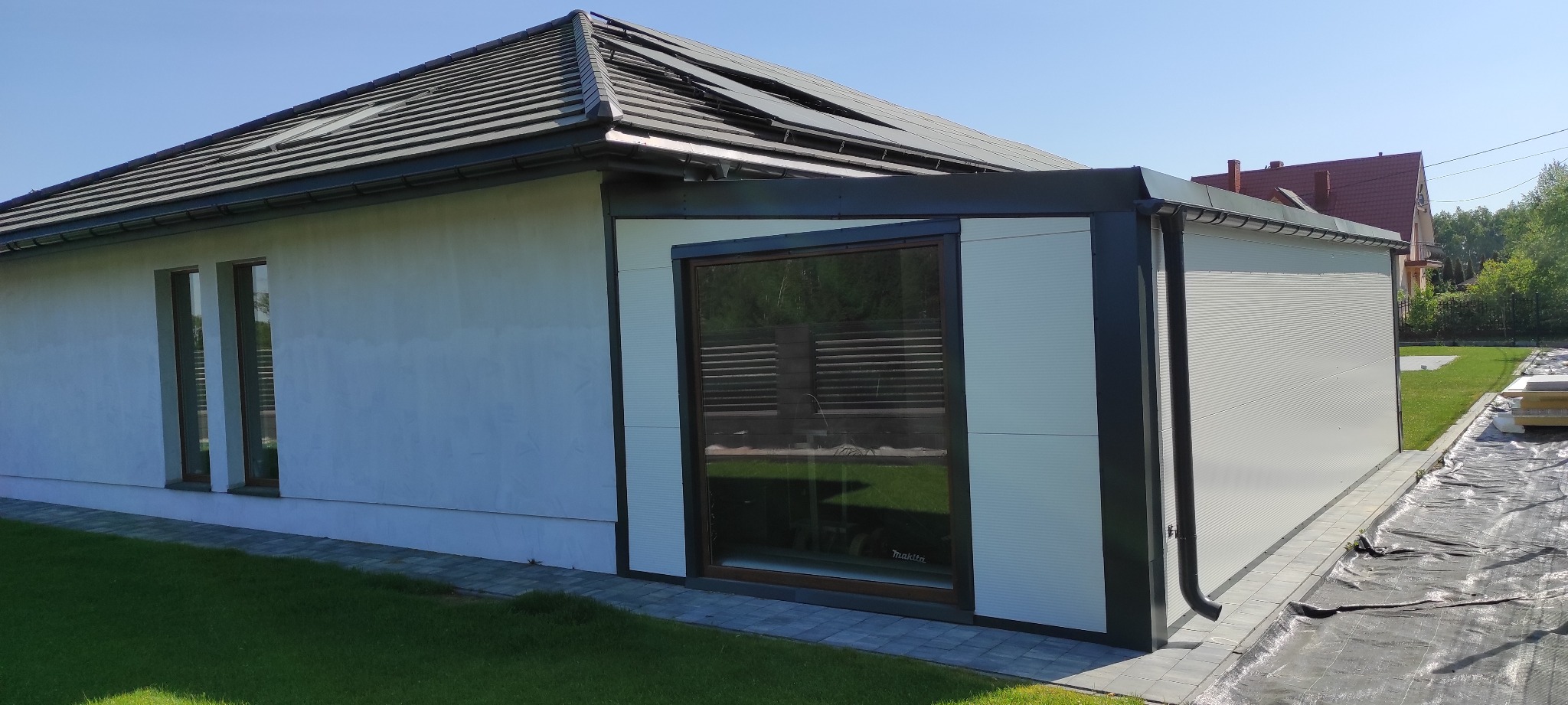 Nowoczesna, biała hala z płyt warstwowych z czarnymi obramowaniami okien i dachu, widoczna z zewnątrz w słoneczny dzień, z ułożoną kostką brukową i przygotowanymi płytami warstwowymi na gruncie.