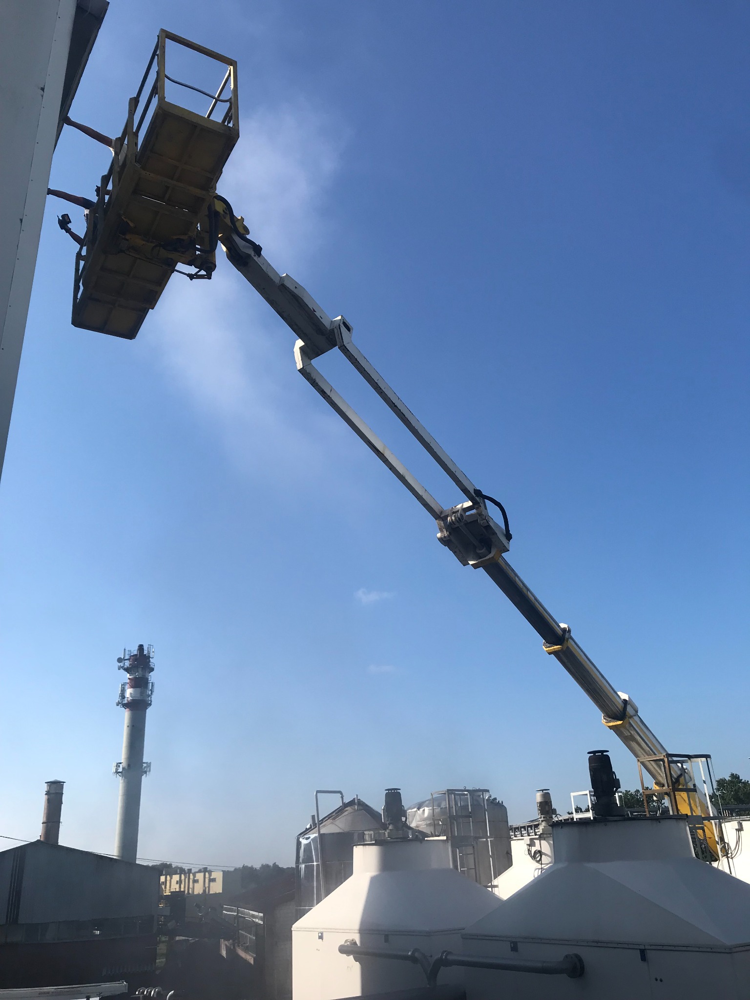 Żółty podnośnik koszowy z dwoma pracownikami przy ścianie budynku przemysłowego, w tle widoczne zbiorniki i komin, błękitne niebo.