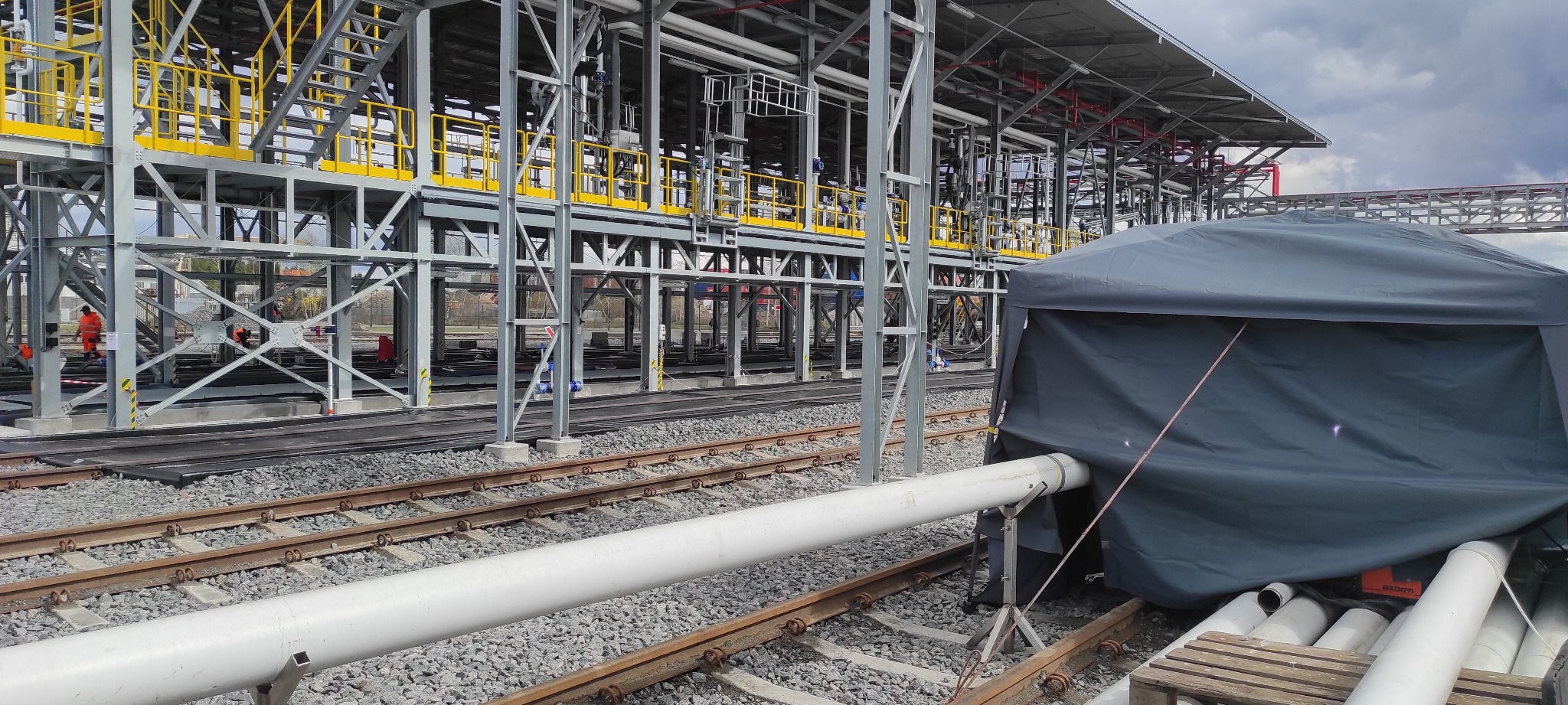 Konstrukcja stalowa z żółtymi barierkami w tle, rury i szary namiot z przodu, widok na plac budowy z torami kolejowymi.