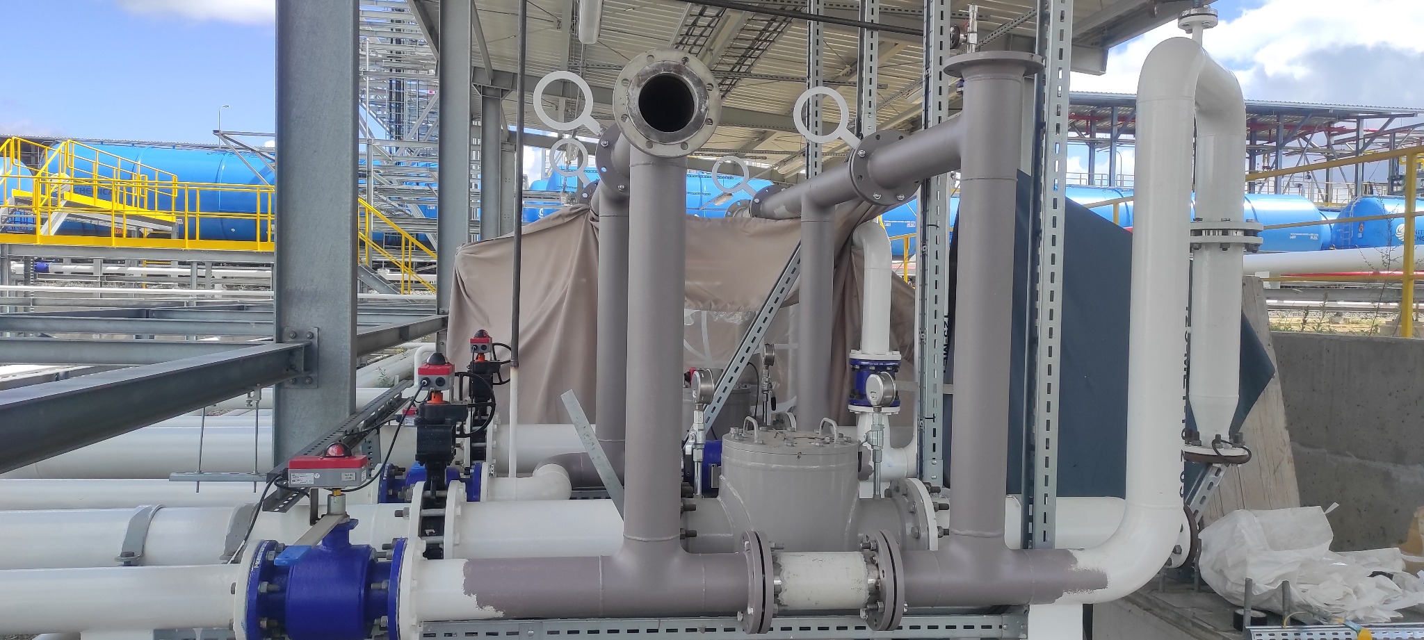 Kompleksowy system rurociągów przemysłowych z licznymi zaworami, manometrami i rurami o różnych średnicach, częściowo osłonięty brezentem, na tle konstrukcji stalowej i zbiorników.