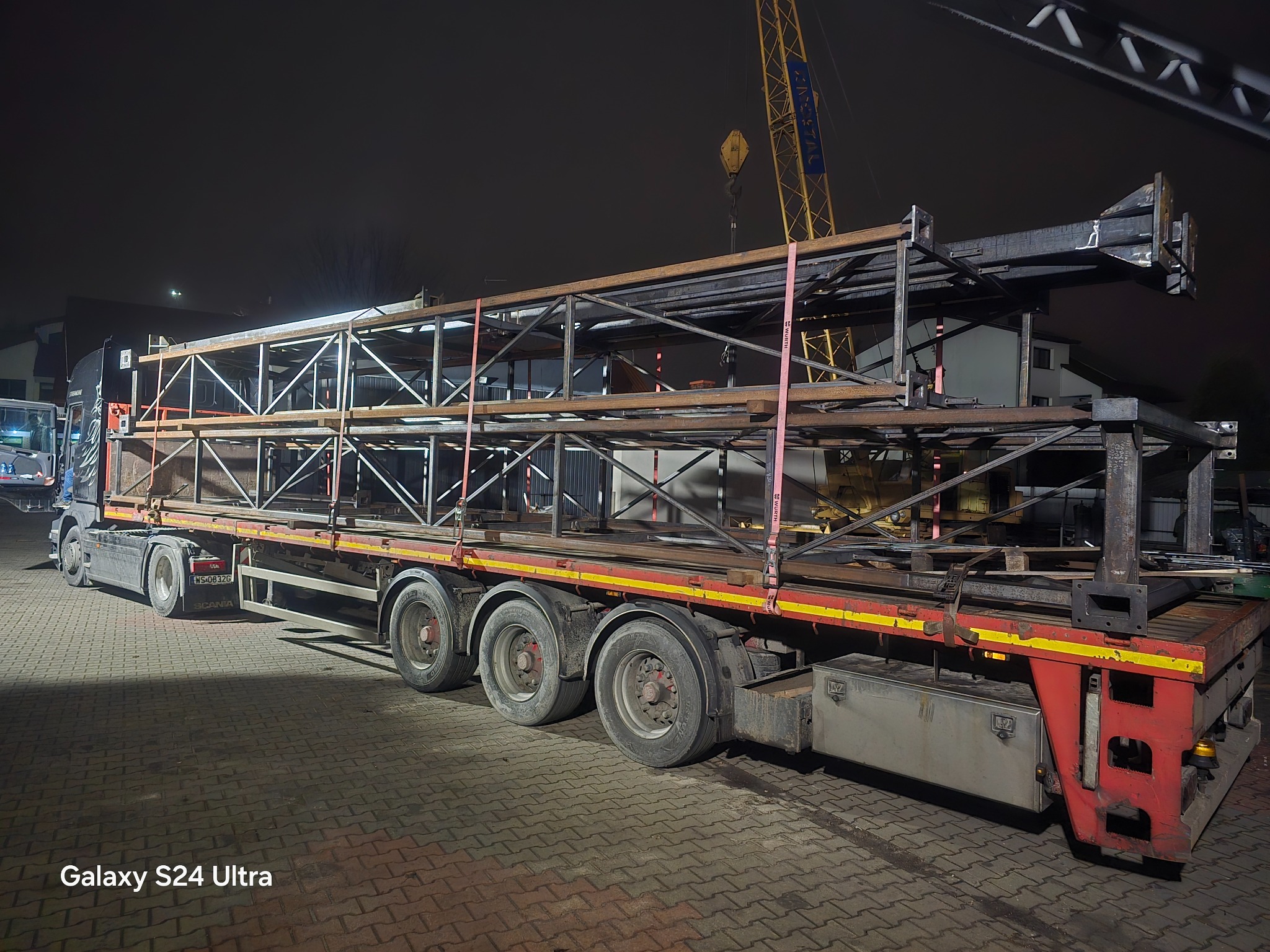 Ciężarówka z naczepą przewożąca stalową konstrukcję kratownicową w nocnych warunkach. Widoczny żuraw w tle.